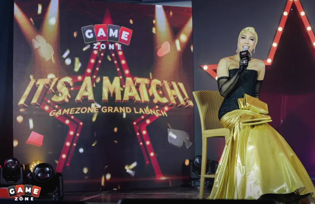 A host in a gold gown speaks on stage at the GameZone "It's a Match!" grand launch