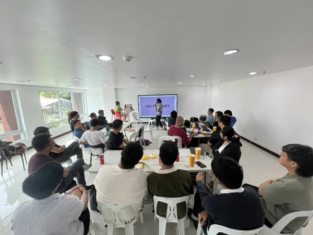 Participants seated at tables watch a facilitator beside a screen reading Pictionary during an empathy training session