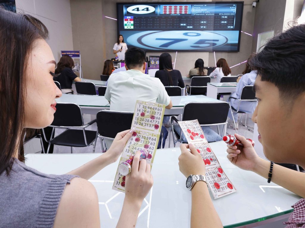 Players marking bingo cards with daubers at a BingoPlus branch while a host calls the numbers on screen