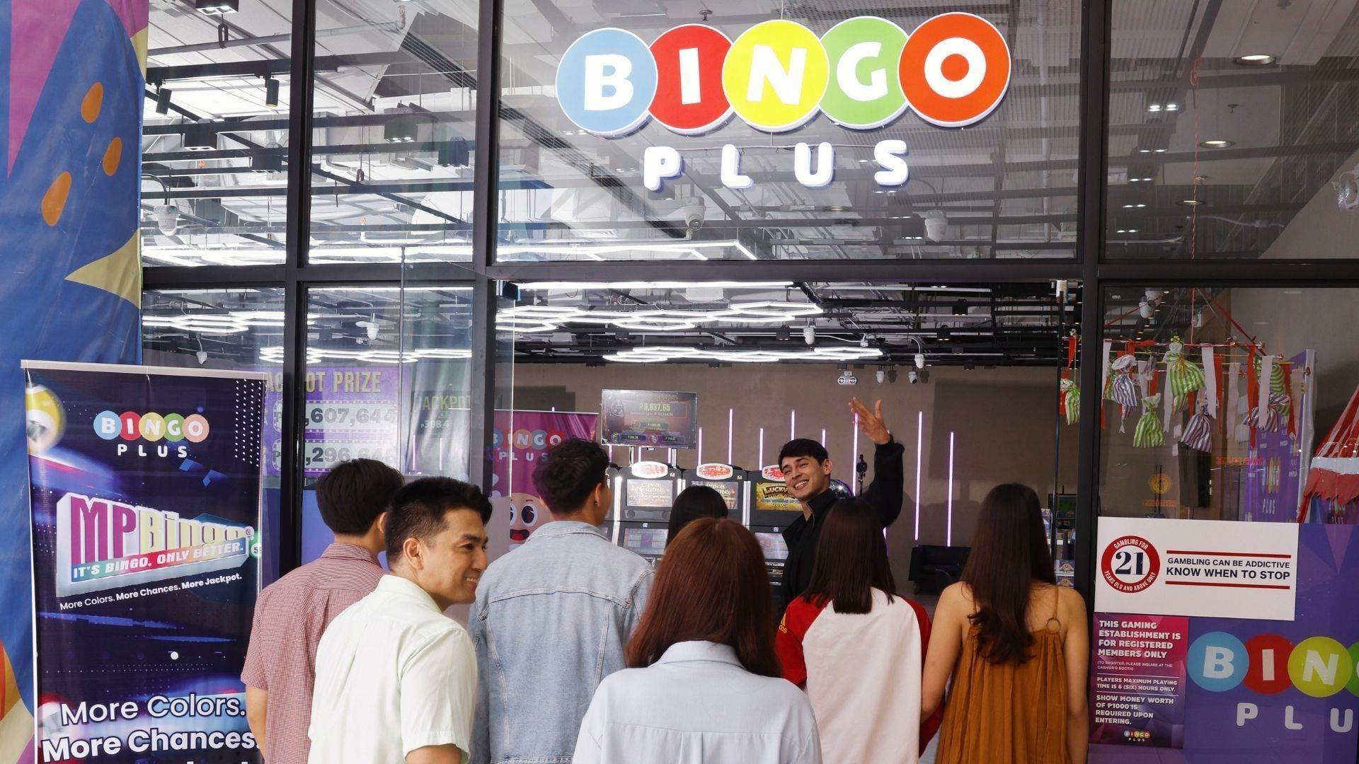 Visitors gather at the glass entrance of a BingoPlus branch as a staff member welcomes them inside