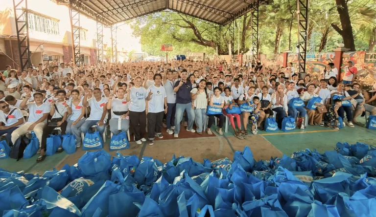 Crowd of relief recipients cheering under a covered court behind rows of DigiPlus Foundation relief bags